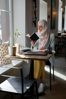 The Edge A woman in a hijab sits at a table, deeply focused on reading a book.