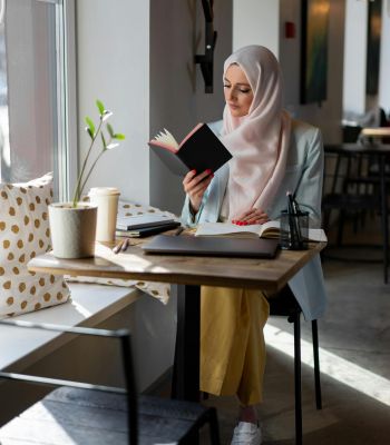 The Edge A woman in a hijab sits at a table, deeply focused on reading a book.