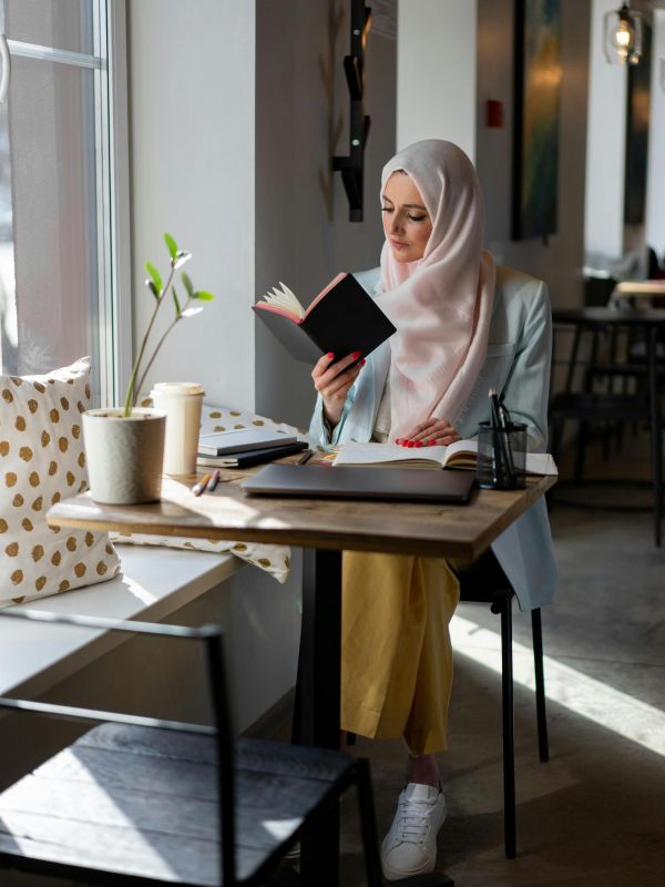 The Edge A woman in a hijab sits at a table, deeply focused on reading a book.